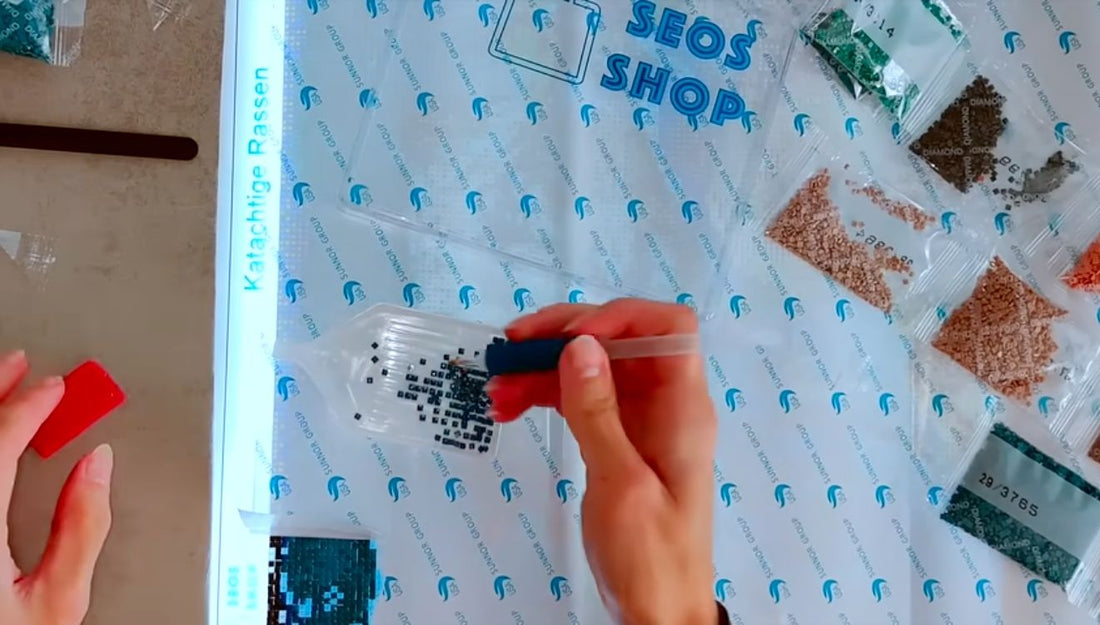 Hand met een diamond-paintingpen boven zwarte steentjes in een wit schudbakje